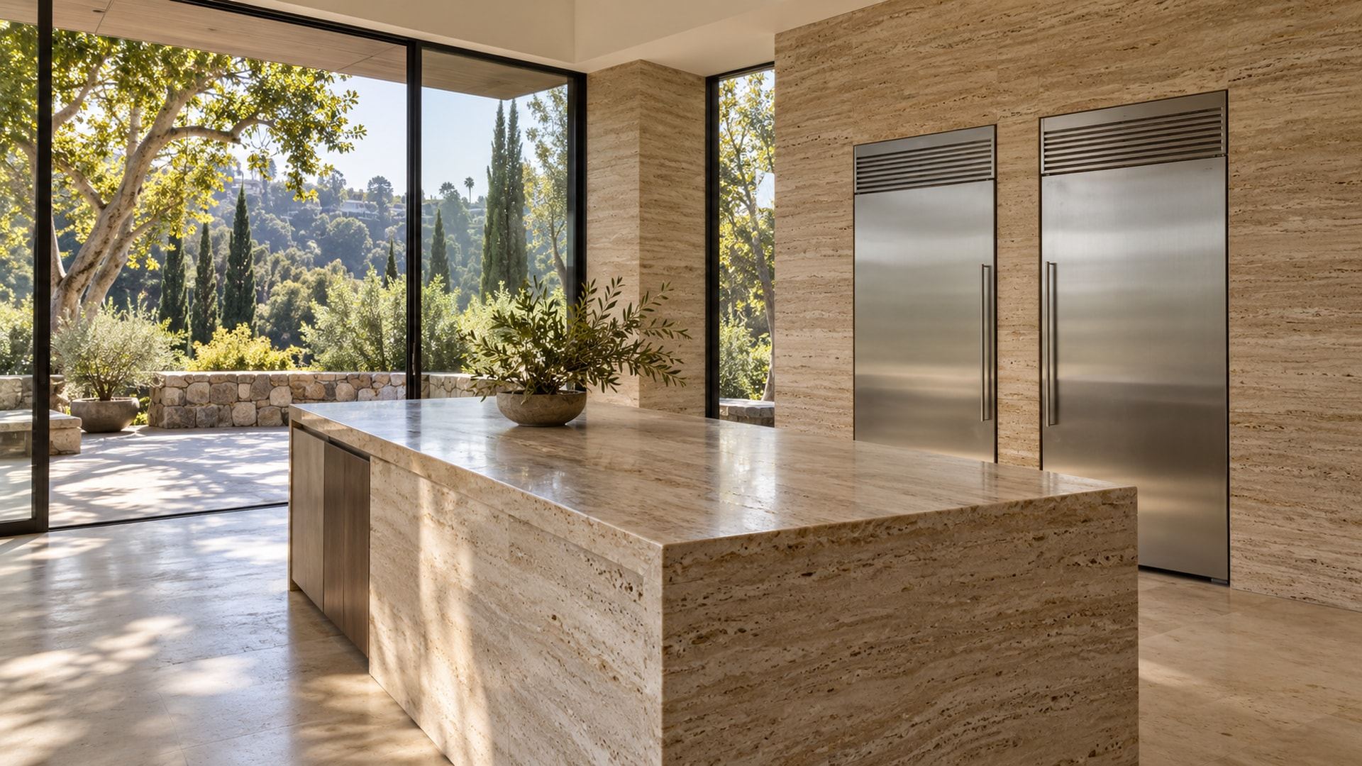 A hillside Bel Air kitchen with two stainless Sub-Zero columns in a travertine wall and a long travertine island, tall windows onto cypress, sycamores and a stone terrace on a green slope